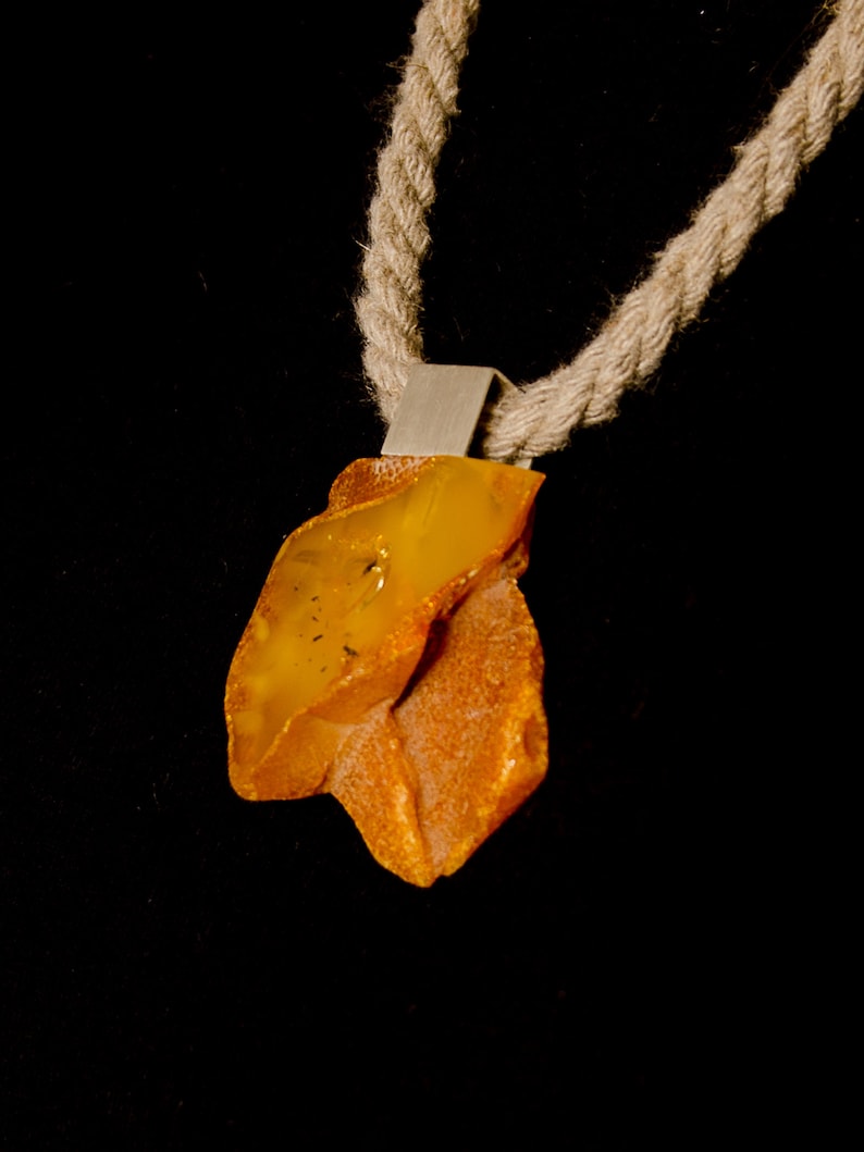Pode incluir: Um colar com um pingente de &acirc;mbar de forma irregular, engastado em prata. O colar &eacute; feito com um cord&atilde;o grosso de corda natural.