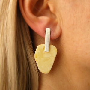 May include: A drop earring featuring a vertical brushed silver bar attached to an irregular, smooth, cream-colored stone pendant. The silver bar extends upward to form the post, while the pale, opaque stone hangs below with a mottled texture.