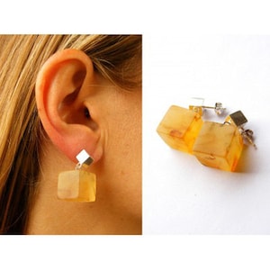 Puede incluir: Un par de pendientes de cubo de ámbar con ganchos de plata. Los pendientes son de color ámbar claro y tienen una apariencia ligeramente translúcida.