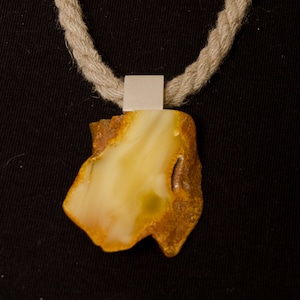 Pode incluir: Um pingente de &acirc;mbar amarelo em um colar de corda. O pingente est&aacute; preso a uma argola de prata.