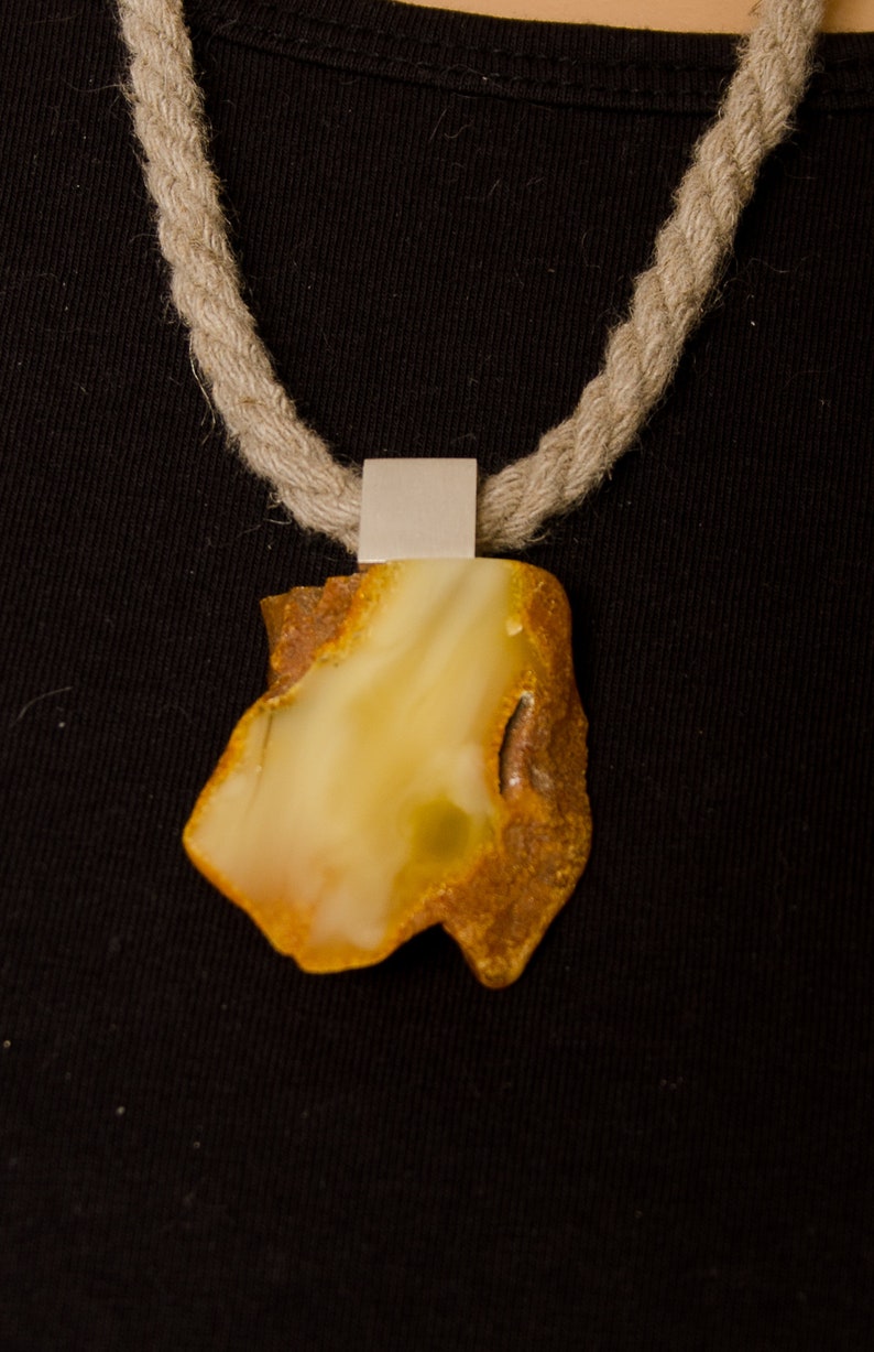 Pode incluir: Um pingente de &acirc;mbar amarelo em um colar de corda. O pingente est&aacute; preso a uma argola de prata.