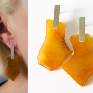 Puede incluir: Un par de pendientes de ámbar con detalles plateados. El ámbar es de un color miel cálido y los pendientes tienen una forma asimétrica única.