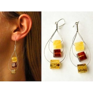 Puede incluir: Un par de pendientes de plata con tres piedras de ámbar en cada pendiente. Las piedras son de color amarillo claro, ámbar oscuro y ámbar claro.