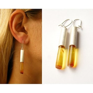 Puede incluir: Un par de pendientes de plata con piedras de ámbar. Los pendientes son largos y delgados, con forma cilíndrica. Las piedras de ámbar son de un color miel cálido.
