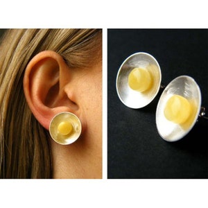 Puede incluir: Un par de pendientes de plata con piedras de ámbar amarillo engastadas en el centro de cada pendiente. Los pendientes son redondos y tienen un borde ligeramente elevado.