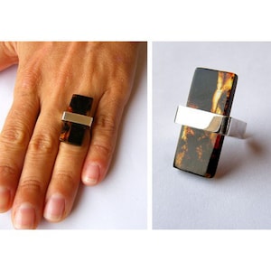 May include: A silver ring featuring a rectangular amber stone. The stone has a brown and black marbled pattern.