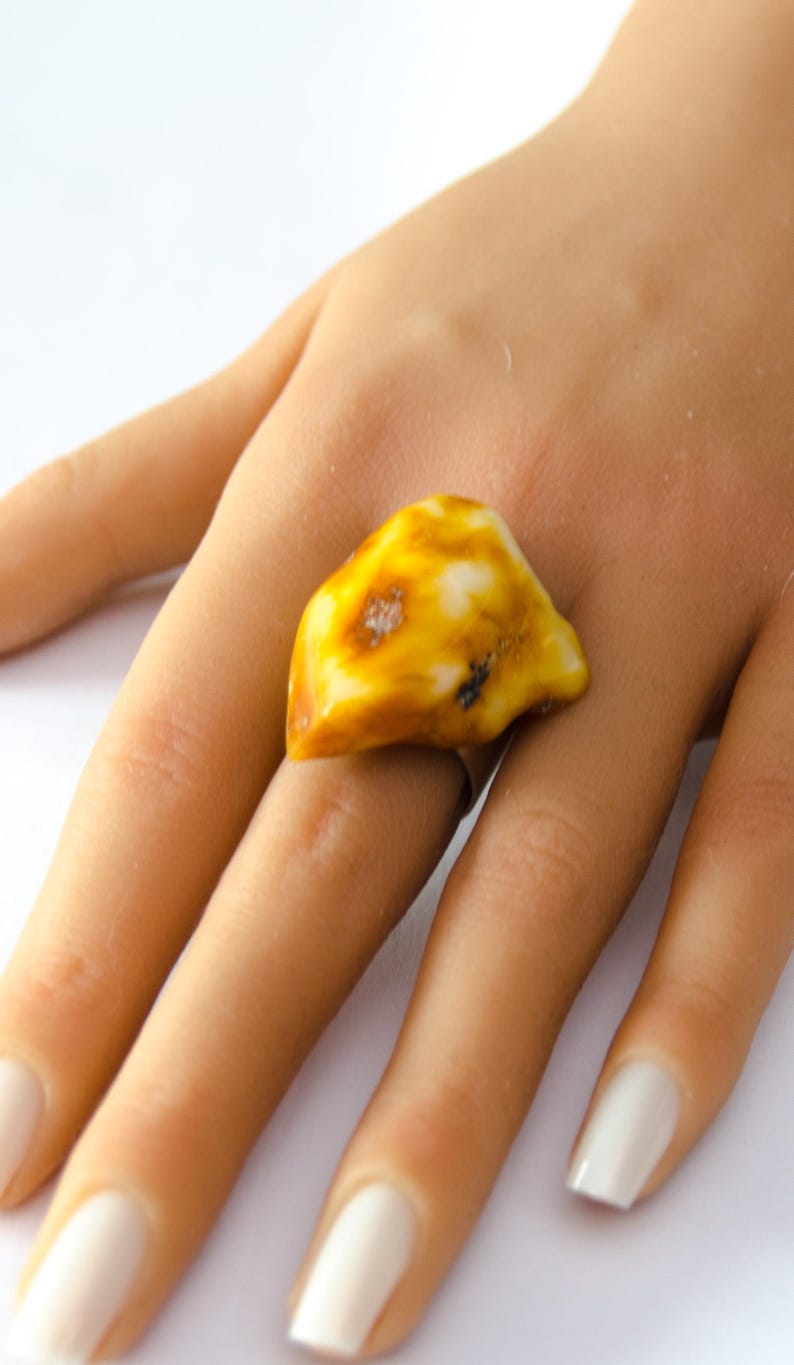 May include: A unique ring featuring a large, irregularly shaped amber stone. The amber is a mix of yellow and white hues, with some darker inclusions. The ring is set on a silver band, creating a striking contrast. This piece is a statement jewelry item.