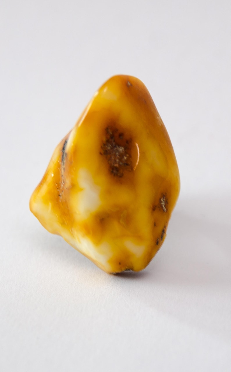 May include: A polished amber gemstone with a smooth, rounded shape. The stone features a mix of translucent yellow and creamy white hues, with a darker, speckled inclusion near the center. The amber is a natural, organic material.