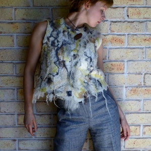 May include: A woman wearing a white, brown, and yellow patterned sleeveless top with a gray pair of pants. She is standing in front of a brick wall.