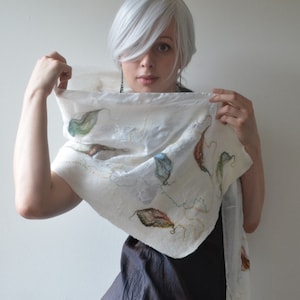 May include: A white silk scarf with embroidered leaves in shades of green, brown, and white. The scarf is draped around a person with white hair.