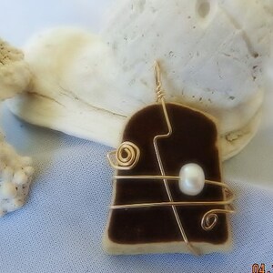 Puede incluir: Un colgante de concha de mar marrón y blanco con alambre dorado envuelto alrededor. Una sola perla blanca está unida al alambre.