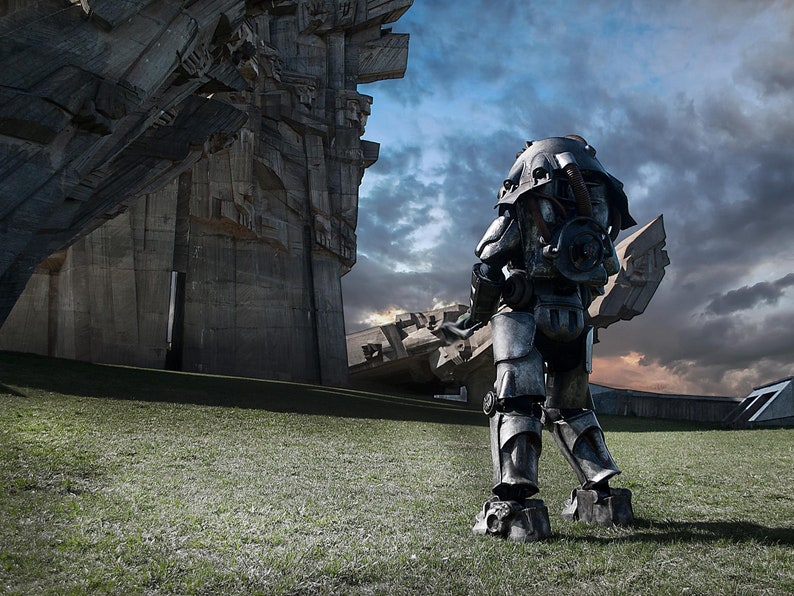 May include: A large, gray, metal robot stands in front of a large, concrete structure. The robot is wearing a helmet and has a large, metal arm. The robot is standing on a grassy field.