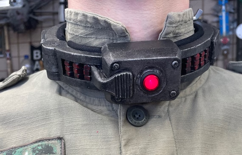 May include: A black and gray metal collar with a red button and a row of red lights. The collar is worn around a person's neck.