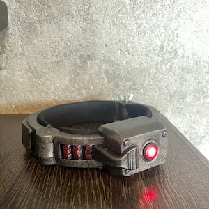 May include: A black and silver futuristic-looking device with a red light. The device has a band that goes around the head and a small red light on the front.