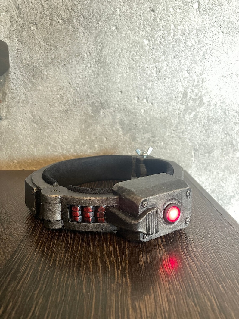 May include: A black and silver futuristic-looking device with a red light. The device has a band that goes around the head and a small red light on the front.