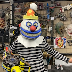 Op de afbeelding: Een wit-zwart gestreept clownsmasker met een blauwe glimlach en een rode neus. Het masker bevindt zich op een witte achtergrond met een verscheidenheid aan andere objecten, waaronder een speelgoedpistool, een speelgoedrobot en een speelgoedauto.