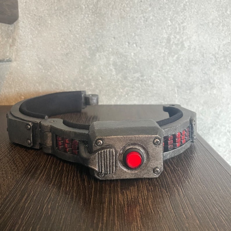 May include: A black and gray headband with a red button and a silver-colored panel. The headband has a textured surface and a series of small red squares.