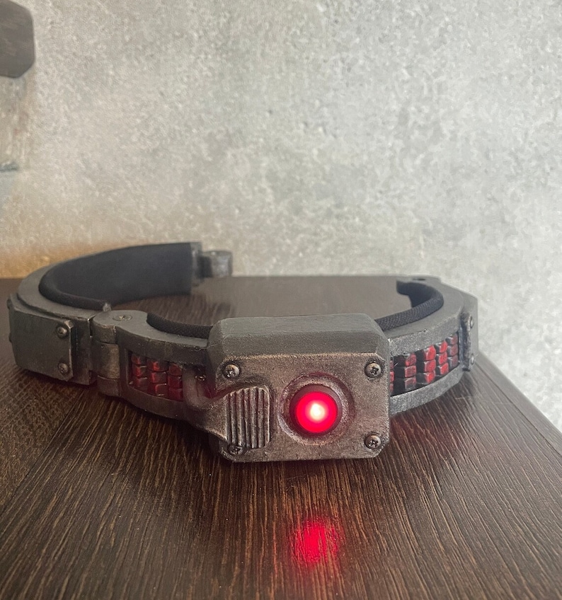 May include: A black and silver headband with a red light in the center. The headband has a textured surface and a series of small, rectangular compartments.