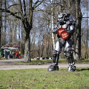 May include: A silver robot wearing a helmet stands in a park. The robot is holding two red circles with the text "Julain Kulps" on them. The robot is standing on a grassy area with trees in the background.