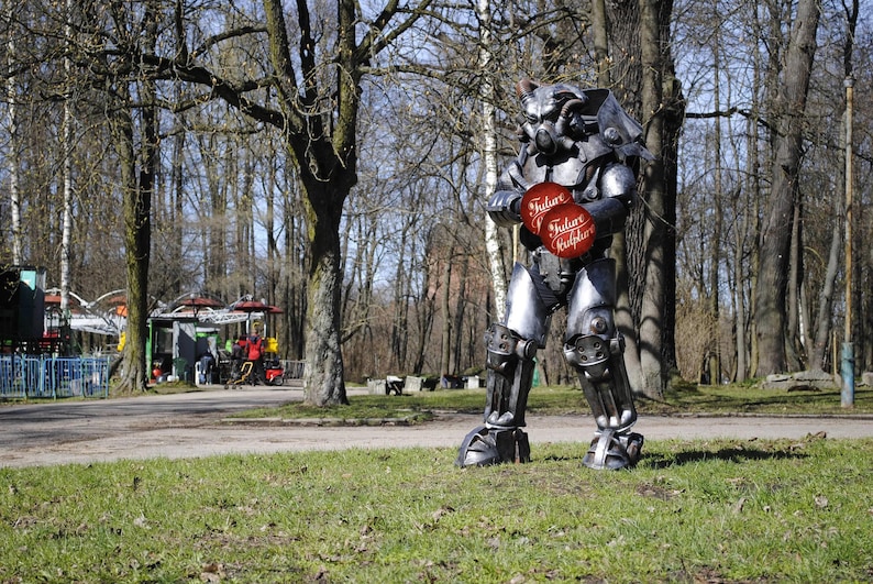 May include: A silver robot wearing a helmet stands in a park. The robot is holding two red circles with the text "Julain Kulps" on them. The robot is standing on a grassy area with trees in the background.