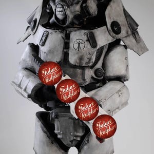 May include: A white and gray robot wearing a helmet with a skull-like face. The robot is holding a weapon with red stickers that say "Future Sculpture" on them.