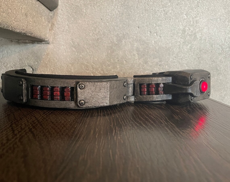 May include: A black and gray headband with a red light on the side. The headband has a series of small, rectangular, red lights on the top.