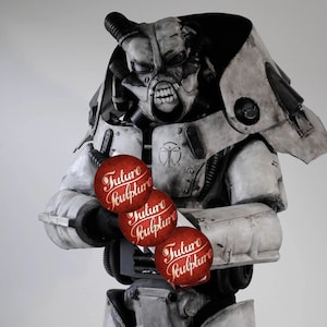 May include: A white and gray robot wearing a helmet with a menacing expression, holding three red spheres with the words "Future Sculpture" written on them.