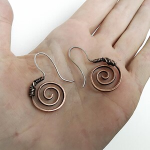 Peut inclure: Une paire de boucles d'oreilles en spirale de couleur cuivre avec des crochets d'oreilles argentés. Le motif en spirale est fait de fil de cuivre, avec les extrémités enveloppées dans un fil plus foncé. Les boucles d'oreilles sont présentées sur un fond clair.