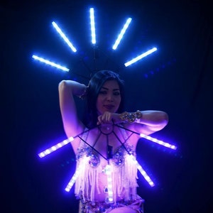 May include: A woman wearing a costume with blue LED lights. The costume includes a headpiece and a bra-like top with fringe. The lights are arranged in a radial pattern, creating a glowing effect against a dark background.