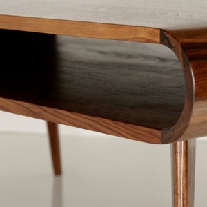 May include: A close-up of a wooden coffee table with a unique curved design. The table has a lower shelf and slender legs.