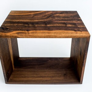 May include: A dark brown wooden cube-shaped end table with a wide, open shelf. The table is made of solid wood with a natural wood grain pattern.