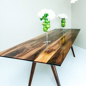 May include: A wooden dining table with a dark brown and light brown wood grain pattern. The table has a glass top and two A-frame legs. Two glass vases with white flowers and green apples are on the table.