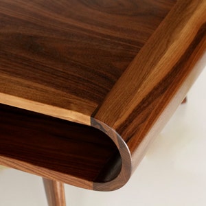 May include: Close-up of a wooden coffee table with a curved edge and a lower shelf. The table is made of dark brown wood with a natural grain pattern.