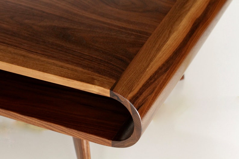 May include: Close-up of a wooden coffee table with a curved edge and a lower shelf. The table is made of dark brown wood with a natural grain pattern.