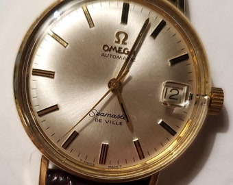 Può includere: Un orologio da polso automatico Omega Seamaster De Ville con cassa rotonda color oro, quadrante color champagne, indici dorati e finestrella della data a ore tre. L'orologio è dotato di un cinturino in pelle marrone scuro.
