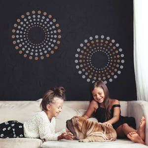 Könnte beinhalten: Zwei dekorative Wandkunstwerke mit konzentrischen Kreisen aus goldenen, silbernen und weißen Punkten vor einer schwarzen, strukturierten Wand. Zwei junge Mädchen sitzen auf einem beigen Sofa und interagieren mit einem Shar Pei Hund.