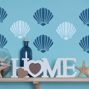 May include: White wooden letters spelling "HOME" with a heart-shaped picture frame and two smaller hearts, on a shelf in front of a blue wall with a seashell stencil design.