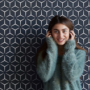 May include: A woman with long brown hair is wearing a light blue fluffy sweater. She is smiling and has her hands on her cheeks. The background is a black and white geometric pattern.