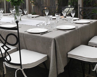 May include: A table set for a dinner party in a backyard. The table is covered with a light gray linen tablecloth and has black metal chairs with white cushions. There are white plates, silverware, and glasses on the table.
