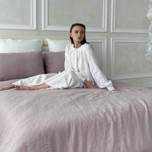 May include: A white linen bedding set with a dusty rose duvet cover and matching pillowcases. A person is sitting on the bed wearing a white linen top.