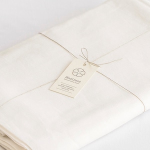 May include: A stack of white linen fabric wrapped with twine and tied with a bow. A tag with the text "BeaLinen Natural Linen" is attached to the twine.