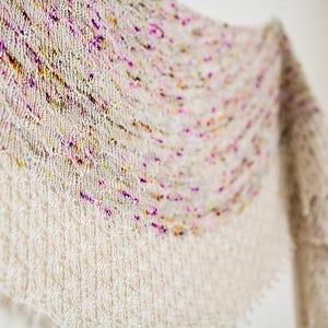 May include: A white knitted shawl with a colourful, speckled pattern. The shawl is draped over a white surface and has a fringed edge.