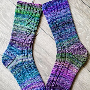 May include: A pair of hand-knitted socks in shades of blue, purple, and green. The socks have a ribbed cuff and a cable knit pattern. They are displayed on metal hangers. The socks are approximately 30 cm long.