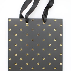 Black gift bag with gold foil cannabis leaf pattern and black ribbon handles.