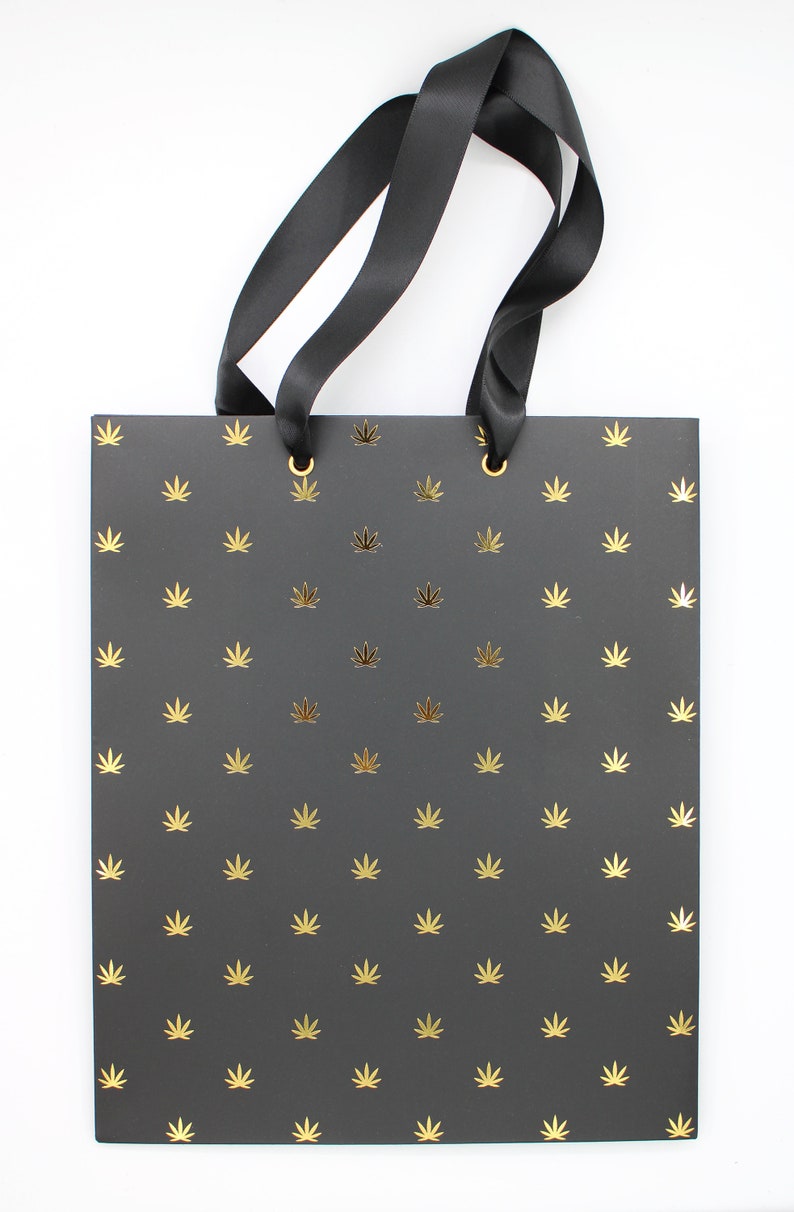 Black gift bag with gold foil cannabis leaf pattern and black ribbon handles.
