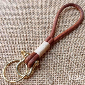May include: A brown leather key chain with a gold metal hook and a loop. The key chain is on a brown burlap background.