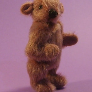 Könnte beinhalten: Ein kleiner, brauner, pelziger Teddybär mit schwarzer Nase und schwarzen Augen. Der Bär steht auf einem violetten Hintergrund.