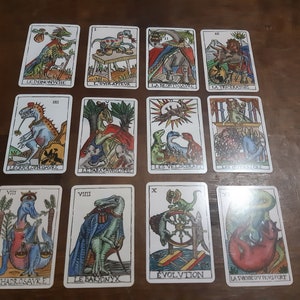 May include: A set of 11 tarot cards featuring colorful illustrations of dinosaurs. The cards are numbered from I to XI and have French text on them. The cards are laid out in three rows on a wooden surface.