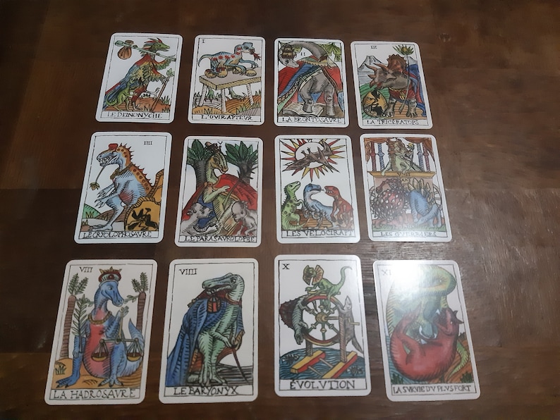 May include: A set of 11 tarot cards featuring colorful illustrations of dinosaurs. The cards are numbered from I to XI and have French text on them. The cards are laid out in three rows on a wooden surface.