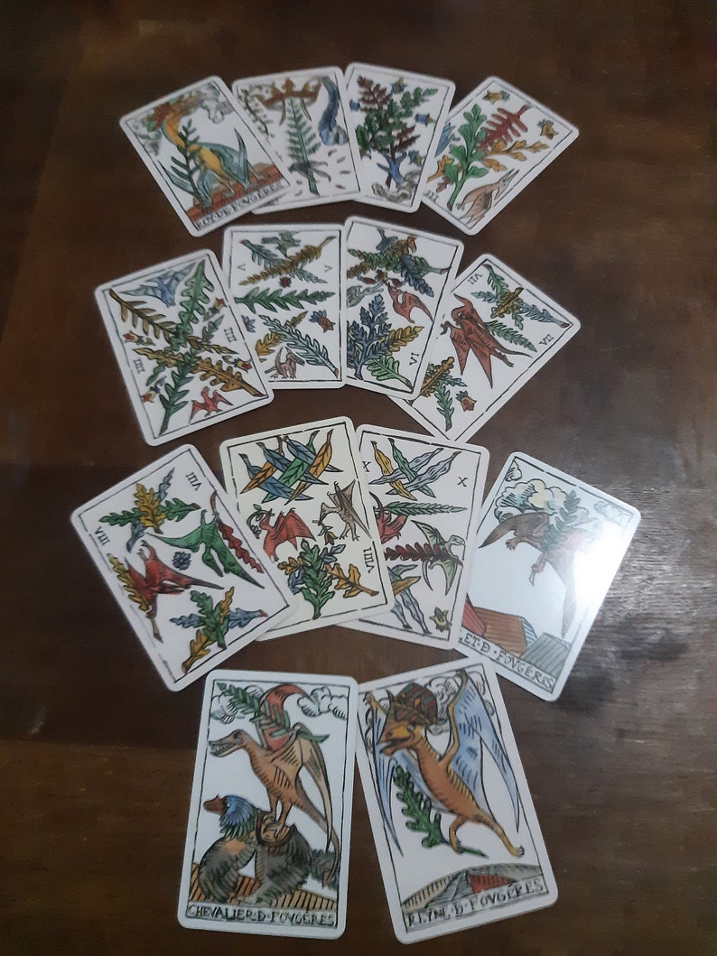 May include: A spread of tarot cards featuring colorful illustrations of winged creatures and foliage. The cards are arranged in a circle on a wooden surface. The cards include the following text: VII, VIII, IX, X, BOY DE FONOL, VIN, CHEVALIER D-FOVGORES, REYM, FOVGERES, ET-D FOVGERIS.